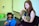 A young woman is playing an acoustic guitar while seated. A young man in a blue shirt and sunglasses holds a microphone, appearing to sing or speak. They are in an indoor setting with a whiteboard or projection screen in the background.