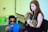 A young woman is playing an acoustic guitar while seated. A young man in a blue shirt and sunglasses holds a microphone, appearing to sing or speak. They are in an indoor setting with a whiteboard or projection screen in the background.