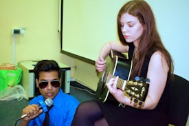 A young woman is playing an acoustic guitar while seated. A young man in a blue shirt and sunglasses holds a microphone, appearing to sing or speak. They are in an indoor setting with a whiteboard or projection screen in the background.