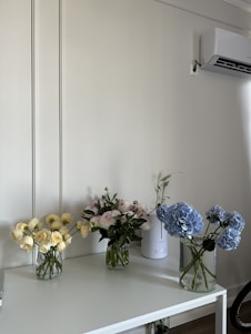 A minimalist interior setting featuring a white table with three glass vases filled with bouquets of flowers. The left vase contains pale yellow roses, the middle vase has light pink carnations, and the right vase holds blue hydrangeas. An air conditioner is mounted on the wall above, and a tall white device, possibly a humidifier or diffuser, sits on the table.