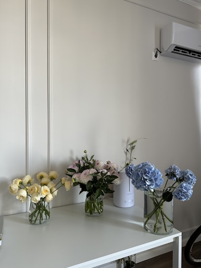 A minimalist interior setting featuring a white table with three glass vases filled with bouquets of flowers. The left vase contains pale yellow roses, the middle vase has light pink carnations, and the right vase holds blue hydrangeas. An air conditioner is mounted on the wall above, and a tall white device, possibly a humidifier or diffuser, sits on the table.