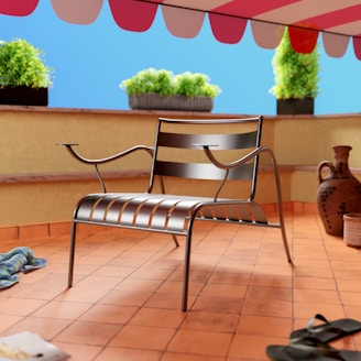 A metallic chair with slatted design is centered on a terracotta tiled patio. The setting is outdoors under a red-and-white striped awning. Three potted plants with lush green foliage sit on a ledge in the background against a bright blue sky. Additional objects include a pair of sandals on the ground, a patterned towel, and clay pots near the chair.