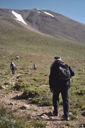 A group of hikers, with one prominent figure in the foreground wearing a backpack, ascends a rocky mountain path. The terrain is covered with sparse vegetation and patches of grass, leading up to a steep, bare mountain slope with some snow patches visible on top. The sky is clear and blue, suggesting a bright, sunny day.