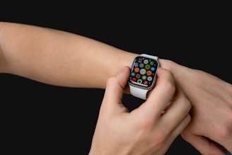 A person interacts with a smartwatch on their wrist, featuring a colorful display of various app icons. The watch has a white band, and the background is solid black.