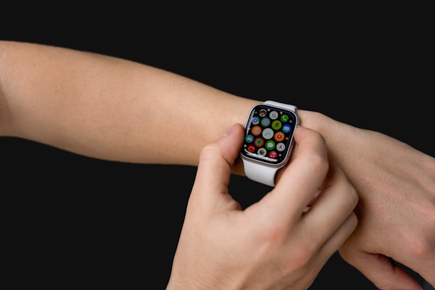 A person interacts with a smartwatch on their wrist, featuring a colorful display of various app icons. The watch has a white band, and the background is solid black.