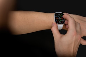 A person interacts with a smartwatch on their wrist, using a finger to select an app on the watch's touchscreen. The smartwatch has a white strap and a black, illuminated display showcasing multiple colorful app icons. The background is dark, highlighting the focus on the device and hand.