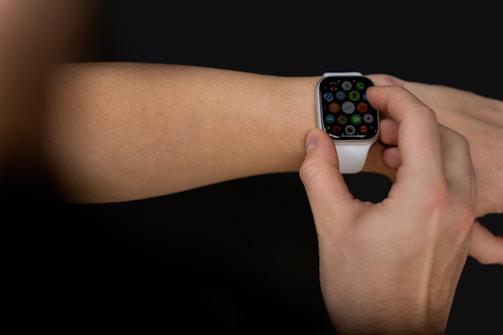 A person interacts with a smartwatch on their wrist, using a finger to select an app on the watch's touchscreen. The smartwatch has a white strap and a black, illuminated display showcasing multiple colorful app icons. The background is dark, highlighting the focus on the device and hand.