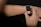 A person interacts with a smartwatch on their wrist, using a finger to select an app on the watch's touchscreen. The smartwatch has a white strap and a black, illuminated display showcasing multiple colorful app icons. The background is dark, highlighting the focus on the device and hand.
