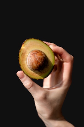 A hand holds half of an avocado against a dark background. The avocado has a large, round seed in the center, surrounded by a creamy, light green flesh. The lighting highlights the texture of the avocado and the skin of the hand, creating a striking visual contrast.
