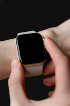 A person wearing a smartwatch on their wrist, with fingers touching the device's screen. The smartwatch features a rectangular face and a light-colored band against a dark background.