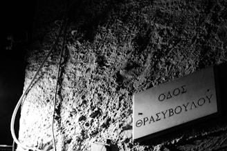 A textured wall with a Greek street sign displaying 'Οδός Θρασυβούλου' in a dimly lit setting. There are wires hanging and the lighting casts shadows that accentuate the roughness of the wall.