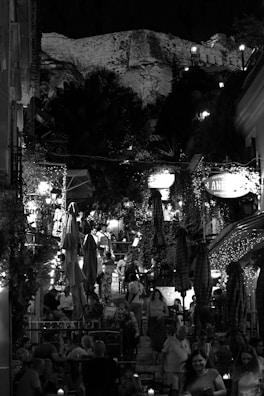A lively and bustling café scene at night with many people sitting and walking around. The area is adorned with lights and umbrellas, while a historic stone structure looms in the background. Lush greenery and vines add to the ambiance, creating an inviting atmosphere.
