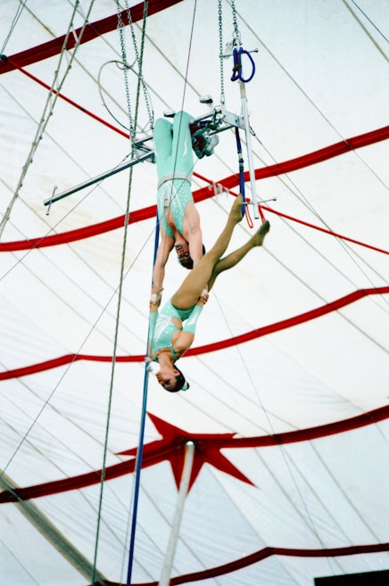 Two acrobats are performing on a trapeze high above the ground. One acrobat hangs upside down, gripping a trapeze bar with her legs. The performers are inside a circus tent, featuring red and white stripes and a large star design on the ceiling.