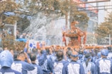 A large group of people dressed in coordinated blue clothing are participating in a traditional festival. They are carrying a highly decorated portable shrine, or mikoshi, while water is being splashed onto them. The scene is taking place in an urban setting with trees and buildings in the background.