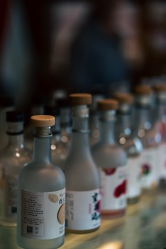 A row of glass bottles with labels and beige caps is lined up on a reflective surface. The bottles are mostly clear or light-colored, and the labels feature various designs and text. The background is blurred, creating a soft focus effect that highlights the bottles in the foreground.