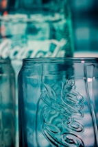 A close-up view of a clear glass bottle with embossed lettering and a textured surface, reflecting a bluish hue from the background lighting.