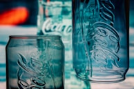 Three empty glass containers with prominent embossed logos stand against a blurred background. The containers are made of clear glass, with reflections and light giving them a bluish tint.