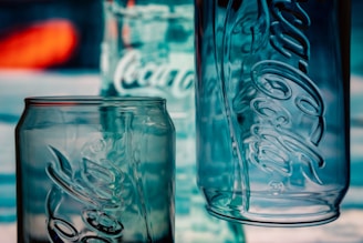 Three empty glass containers with prominent embossed logos stand against a blurred background. The containers are made of clear glass, with reflections and light giving them a bluish tint.
