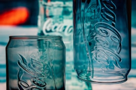 Three empty glass containers with prominent embossed logos stand against a blurred background. The containers are made of clear glass, with reflections and light giving them a bluish tint.