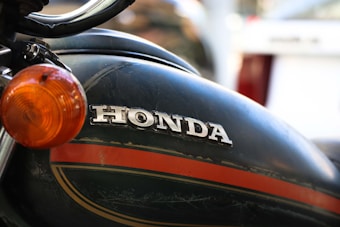 A close-up view of a motorcycle with a visible Honda logo on the fuel tank. The tank features a dark green and orange stripe design. The image also shows a portion of the motorcycle's chrome handlebar and an orange indicator light.