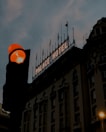 A large hotel building with a prominent 'Palace Hotel' sign illuminated against the darkening sky. The building features ornate architectural details and several flags mounted on the roof. In the foreground, a traffic light shows an amber light, casting a warm glow.