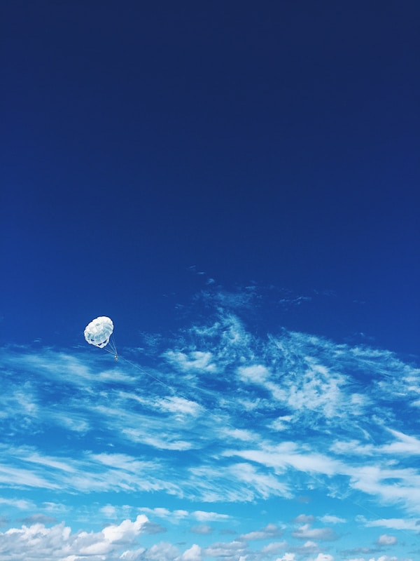 A bright blue sky with scattered white clouds dominates the scene. A single parachute with a person attached floats gently among the clouds, creating a sense of tranquility and freedom.