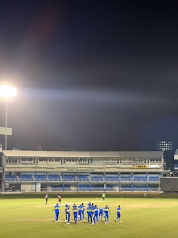 A group of athletes in blue uniforms stands on a sports field under bright stadium lights. The stadium seating is visible in the background, and a few players in different colored uniforms stand separately. The night sky is dark, contrasting with the illuminated field.