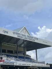 A pavilion at a cricket club is shown with a gabled roof bearing the name 'QUEEN'S PARK CRICKET CLUB BRIAN LARA PAVILION'. A large clock is positioned on the front of the roof. Below, there are multiple levels of blue seating and a balcony marked as 'JOEY CAREW MEMBERS BALCONY'. The structure also features metal railings and supporting beams, with a sign for BP Trinidad and Tobago visible.