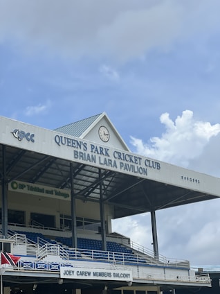A pavilion at a cricket club is shown with a gabled roof bearing the name 'QUEEN'S PARK CRICKET CLUB BRIAN LARA PAVILION'. A large clock is positioned on the front of the roof. Below, there are multiple levels of blue seating and a balcony marked as 'JOEY CAREW MEMBERS BALCONY'. The structure also features metal railings and supporting beams, with a sign for BP Trinidad and Tobago visible.