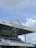 A pavilion at a cricket club is shown with a gabled roof bearing the name 'QUEEN'S PARK CRICKET CLUB BRIAN LARA PAVILION'. A large clock is positioned on the front of the roof. Below, there are multiple levels of blue seating and a balcony marked as 'JOEY CAREW MEMBERS BALCONY'. The structure also features metal railings and supporting beams, with a sign for BP Trinidad and Tobago visible.