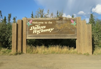 A large wooden sign stands amidst trees and grass, reading 'Welcome to the James Dalton Highway' and 'Gateway to the Arctic - the Road to Prudhoe Bay'. It features images of pink flowers and an outline of Alaska against a backdrop of greenery and a partly cloudy sky.