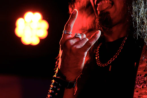 A person is making a hand gesture commonly associated with rock music, known as the 'devil horns.' The background features a bright, blurred, circular light source. The person wears multiple rings, a spiked wristband, and a chain necklace. Their tongue is out, adding to the rock or metal aesthetic.