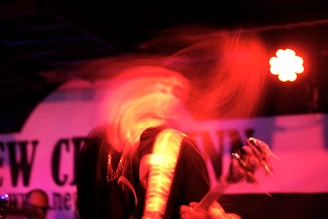 A dynamic and blurred image depicts a musician in motion, possibly a guitarist, under red lighting with a visible guitar. The background contains a partially obscured sign or banner.