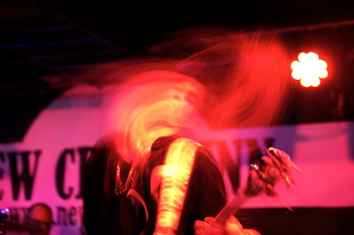 A dynamic and blurred image depicts a musician in motion, possibly a guitarist, under red lighting with a visible guitar. The background contains a partially obscured sign or banner.