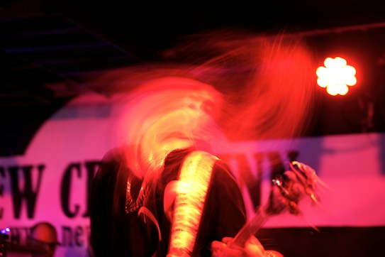 A dynamic and blurred image depicts a musician in motion, possibly a guitarist, under red lighting with a visible guitar. The background contains a partially obscured sign or banner.