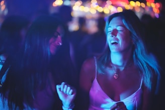 Two women are dancing and enjoying themselves in a dimly lit environment. The background features colorful, blurred lights which add to the lively atmosphere. One woman is wearing a pink top, while the other has long dark hair. Both appear to be smiling and immersed in the moment.