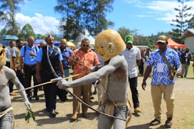 A group of people, some in traditional tribal attire, are gathered outdoors. The central figure is adorned in body paint and a distinctive mask, holding a traditional bow. Others in the scene are wearing modern clothing, including shirts with bright patterns. The setting appears to be a cultural or ceremonial event, with trees and tents visible in the background.