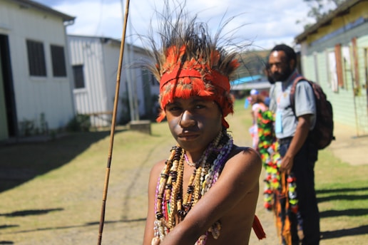 A person wearing traditional attire, including a vibrant red headdress with feathers and an assortment of colorful necklaces. They are in an outdoor setting with buildings in the background. Another person is visible in the distance, dressed casually.