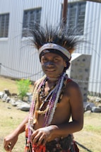 A person is wearing traditional attire, with an elaborate headdress made of feathers and decorative beaded necklaces. The background includes a corrugated metal building and a rocky landscape.
