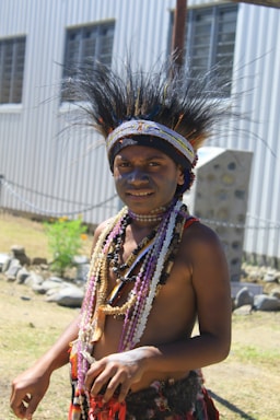 A person is wearing traditional attire, with an elaborate headdress made of feathers and decorative beaded necklaces. The background includes a corrugated metal building and a rocky landscape.