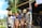 A group of people dressed in traditional tribal attire, with some wearing body paint and elaborate headpieces, pose for a photo. They are standing in front of a building with festive decorations and greenery around them. Bright colored beads and costumes are prominent.