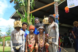 A group of people dressed in traditional tribal attire, with some wearing body paint and elaborate headpieces, pose for a photo. They are standing in front of a building with festive decorations and greenery around them. Bright colored beads and costumes are prominent.
