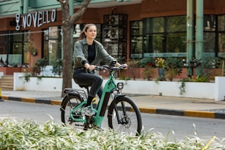 A person rides a green electric bicycle along a street lined with greenery and modern buildings. The background includes a sign for a place called Novello and some trees. The atmosphere is urban and tranquil.