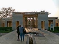 A modern architectural entrance features a symmetric design with light-colored walls and a small fountain in the center of a symmetrical stone-paved walkway. The entrance is surrounded by neatly trimmed greenery and palm trees. Two people are standing in the foreground, engaging in conversation.