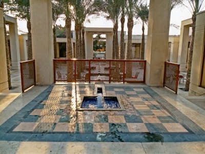 A serene courtyard with a small water fountain at the center, surrounded by palm trees. The space is enclosed by beige columns and a decorative iron fence with geometric patterns. Sunlight casts intricate shadows across a checkered stone floor, creating a tranquil and inviting atmosphere.