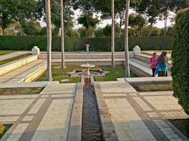 A serene garden setting featuring a symmetrical stone pathway lined with trees and bordered by trimmed hedges. The center holds a small fountain with intricate geometric designs, surrounded by neatly laid out grass and stone benches. Two people walk along the path, engaged in conversation.
