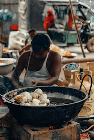A man is sitting and preparing food in a large black metal pan filled with oil, placed over an open flame. The pan contains several round, dough-like items being fried. The background shows a busy street scene with people, one of whom is wearing a red sari, and a vehicle resembling a tuk-tuk.