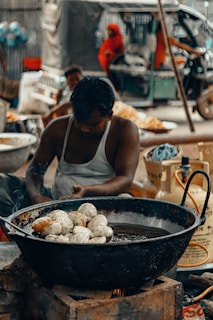 A man is sitting and preparing food in a large black metal pan filled with oil, placed over an open flame. The pan contains several round, dough-like items being fried. The background shows a busy street scene with people, one of whom is wearing a red sari, and a vehicle resembling a tuk-tuk.