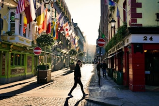 A vibrant street scene featuring a row of colorful buildings adorned with numerous international flags. The cobblestone street is bathed in warm sunlight, creating long shadows. People casually walk along the street, with one individual prominently crossing in the foreground. The scene captures the lively and multicultural atmosphere of the area.