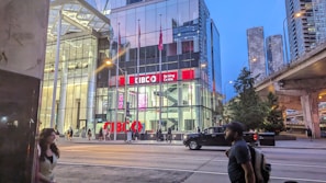 A modern urban scene featuring a glass-fronted building with a sign reading 'CIBC Banking Centre.' The setting is in a city environment with skyscrapers visible in the background. There's a road in front with pedestrians walking and a pickup truck driving by. It's evening or early night, as indicated by the lighting and the sky's color.